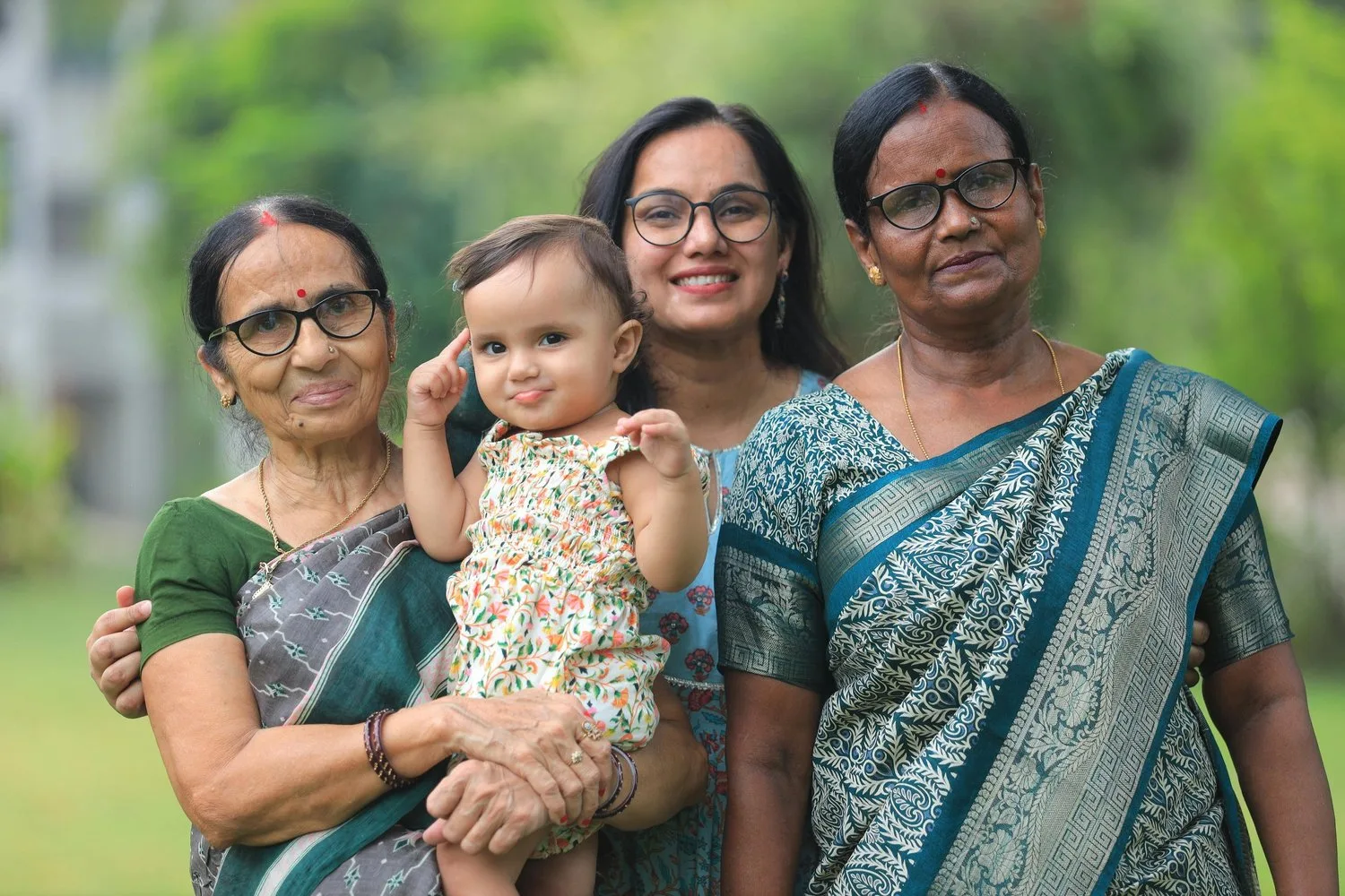 Two older women and a younger woman stand together holding a baby outdoors, all smiling.
