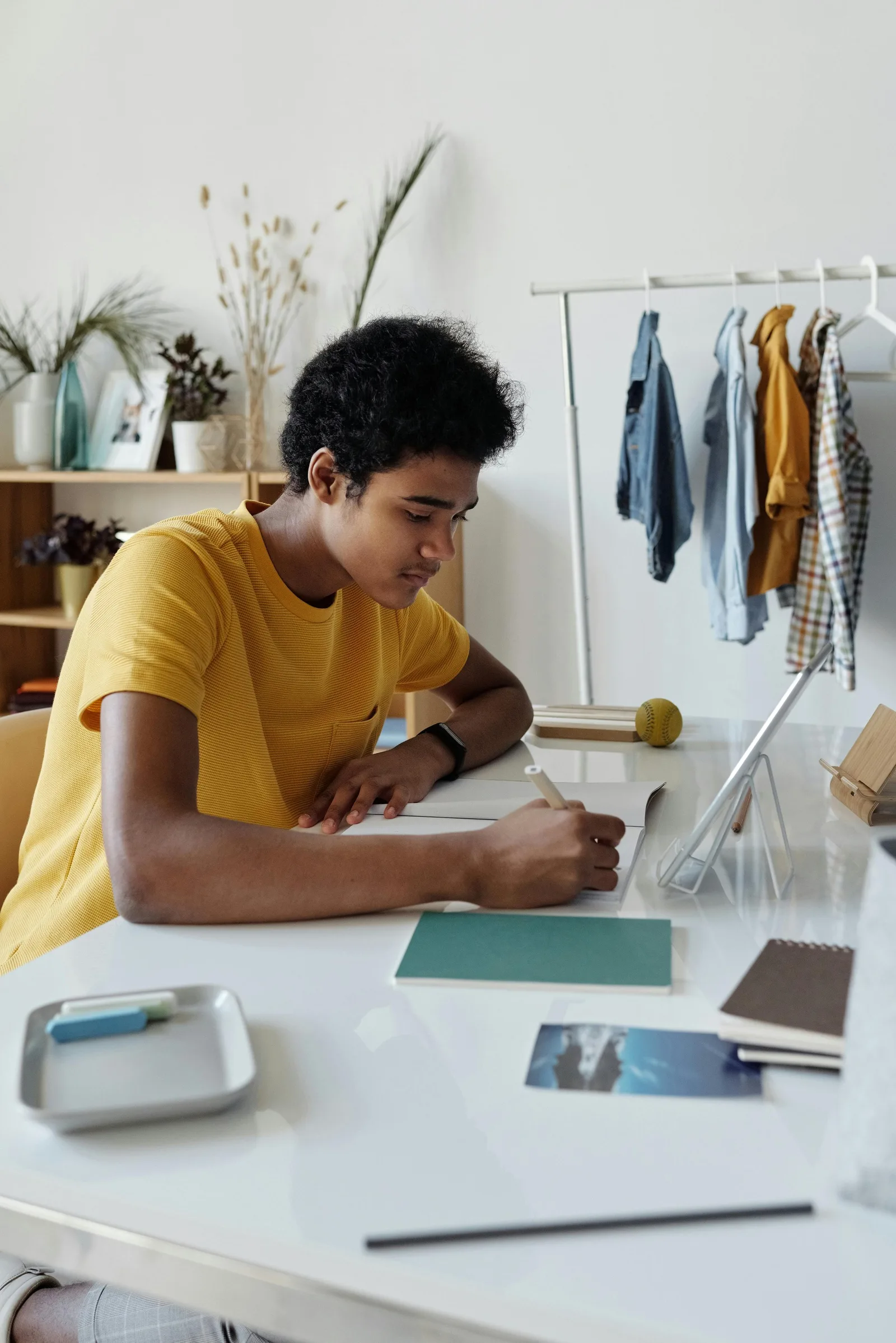A teenager in a yellow shirt writes at a desk near a clothing rack and houseplants.