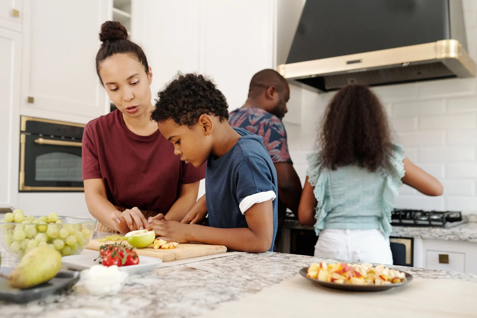 A mother helps a boy cut fruit at a kitchen counter while a father and girl cook in the background.