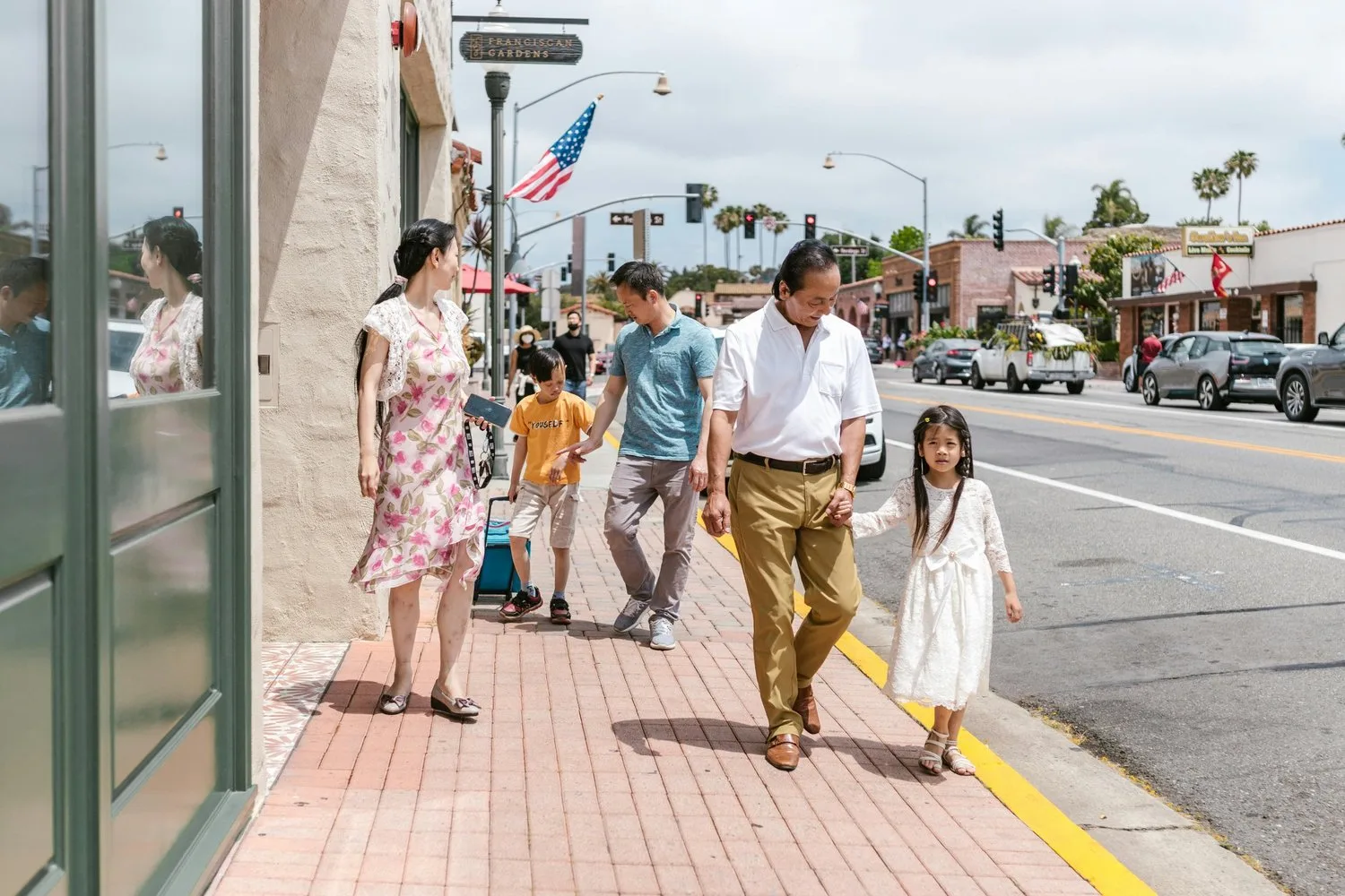 A family of five walks together down a sidewalk on a sunny street holding hands.