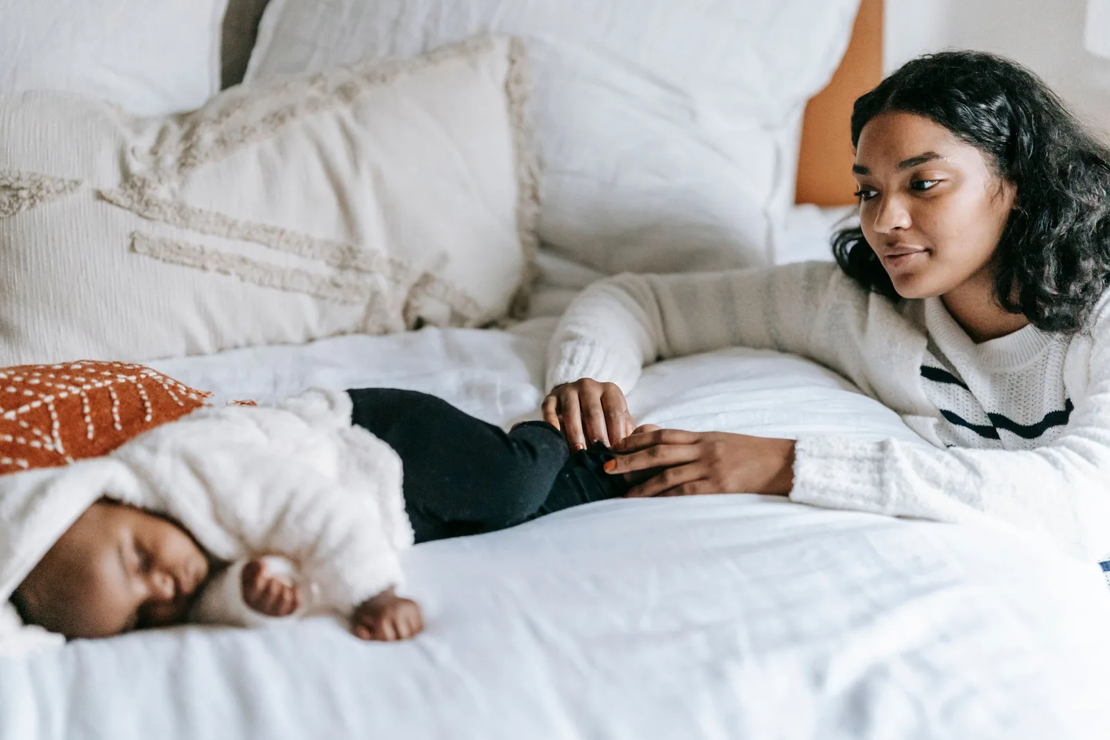 A woman lies in bed watching a sleeping newborn wrapped in a blanket beside her.