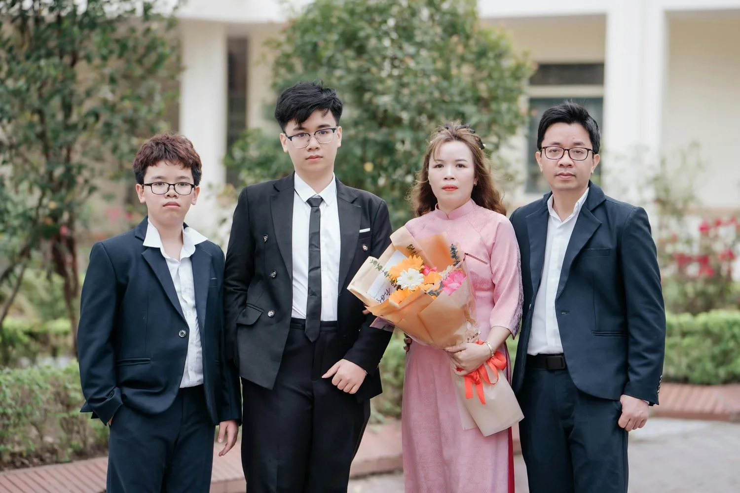 Four people in formal attire stand together outdoors, one woman holding a bouquet of flowers.