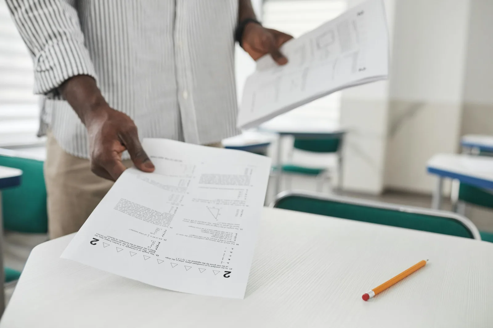 A man stands in a classroom holding worksheets, with more papers and a pencil on a desk in front of him.