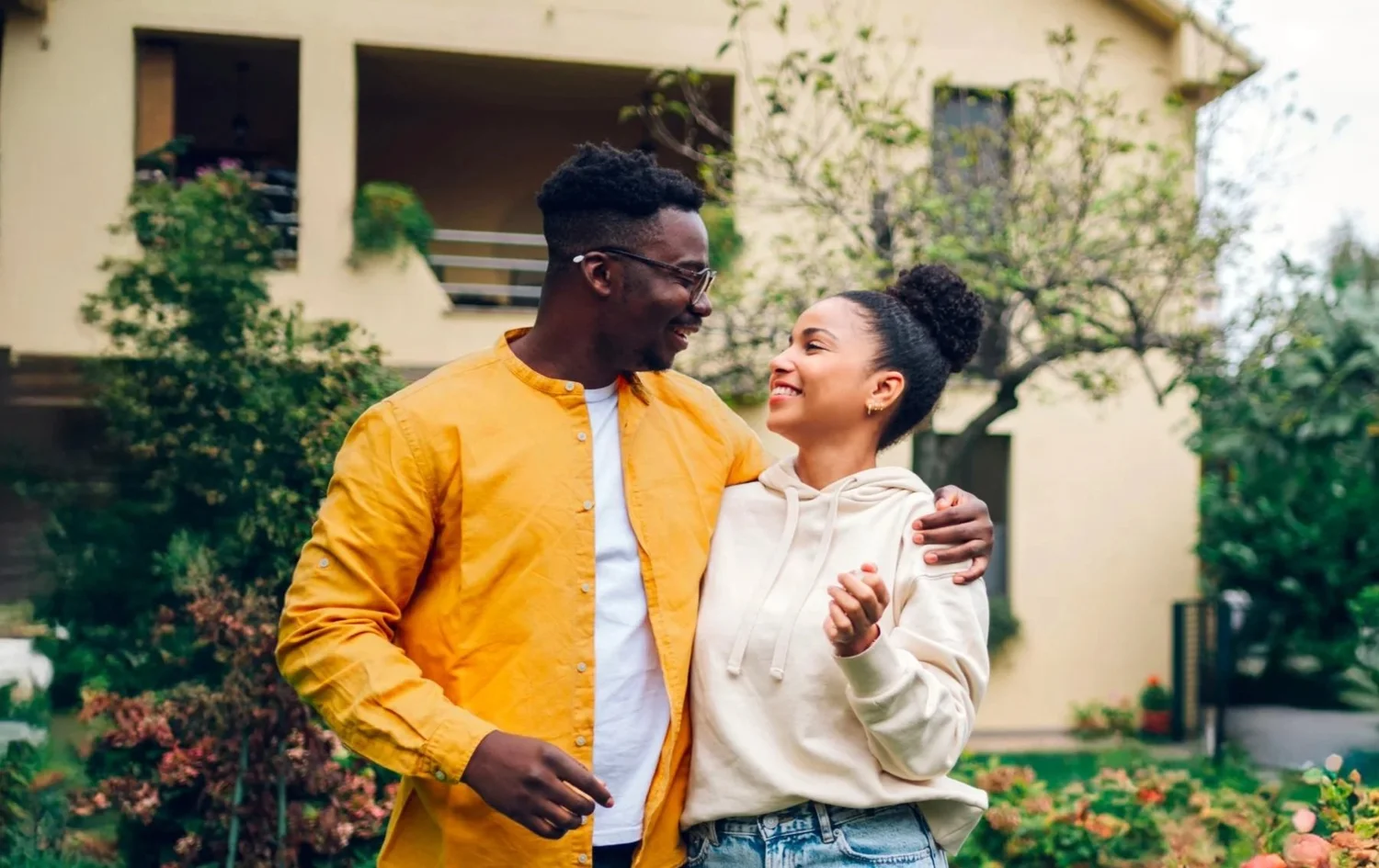 A happy couple embracing outdoors in front of a house with green plants and trees, smiling at each other.
