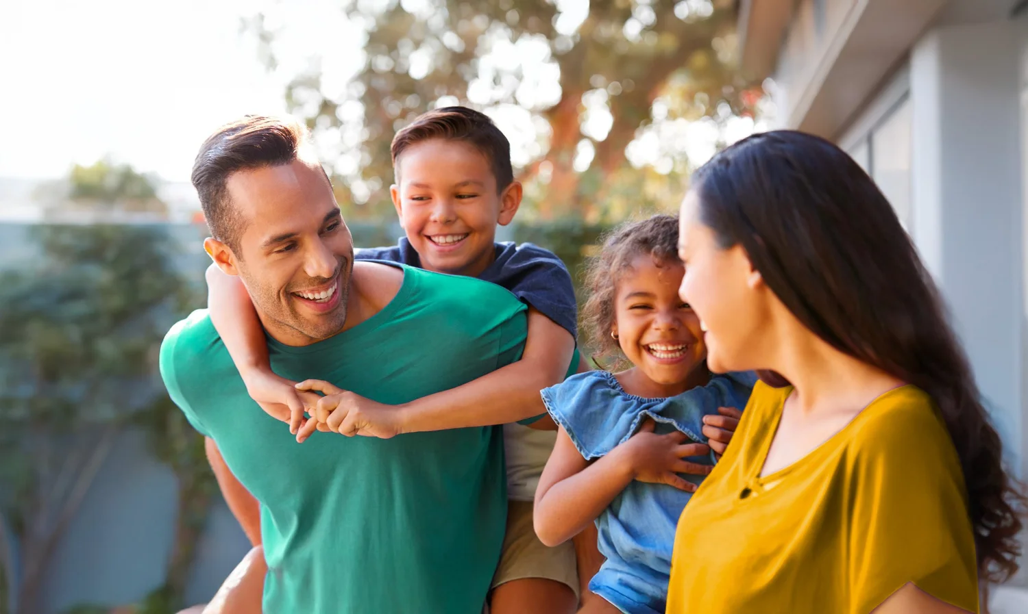 A family of four enjoying time outdoors, with the father giving his son a piggyback ride. The mother is smiling at the father and children, all appearing happy and engaged.