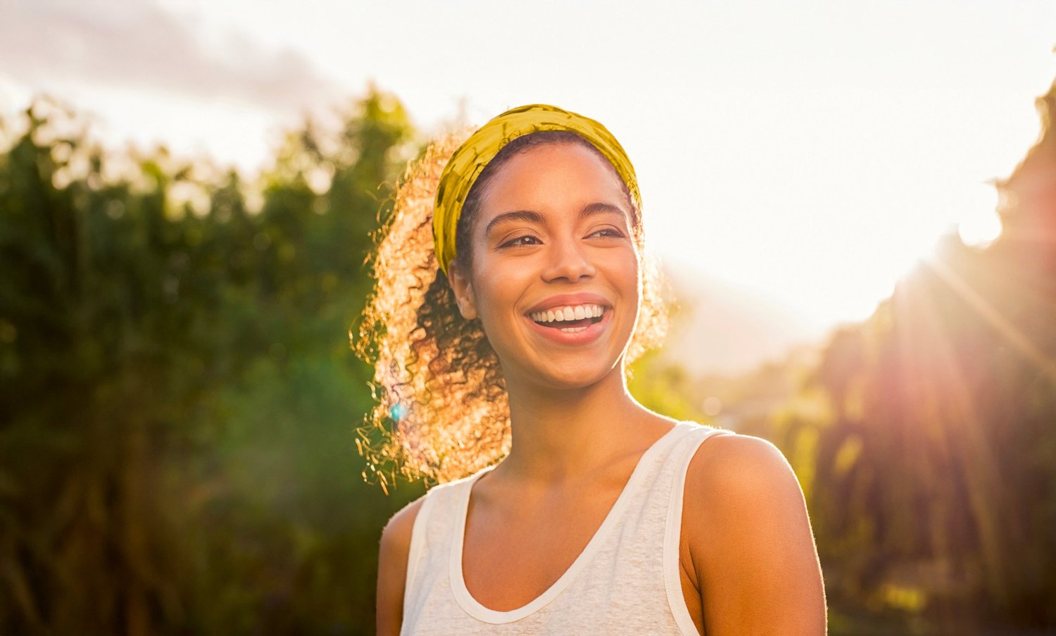 A young woman smiling outdoors in bright sunlight, wearing a yellow headband and a white sleeveless top, with trees in the background.